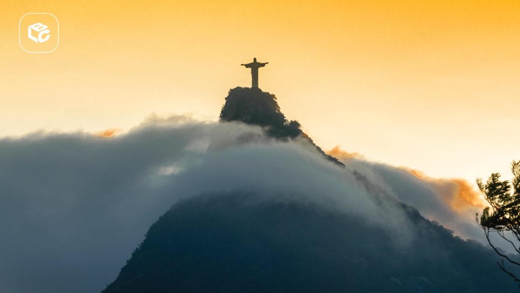 Vista do Cristo Redentor no topo de uma montanha, parcialmente envolto por nuvens densas durante o nascer ou pôr do sol. O monumento aparece em silhueta contra um céu em tons dourados e alaranjados, enquanto a neblina cobre grande parte da encosta. A composição destaca a paisagem natural e o contraste entre a estátua e as nuvens, criando uma cena icônica da cidade do Rio de Janeiro. No canto superior esquerdo há um logotipo branco sobre fundo translúcido.