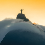Vista do Cristo Redentor no topo de uma montanha, parcialmente envolto por nuvens densas durante o nascer ou pôr do sol. O monumento aparece em silhueta contra um céu em tons dourados e alaranjados, enquanto a neblina cobre grande parte da encosta. A composição destaca a paisagem natural e o contraste entre a estátua e as nuvens, criando uma cena icônica da cidade do Rio de Janeiro. No canto superior esquerdo há um logotipo branco sobre fundo translúcido.